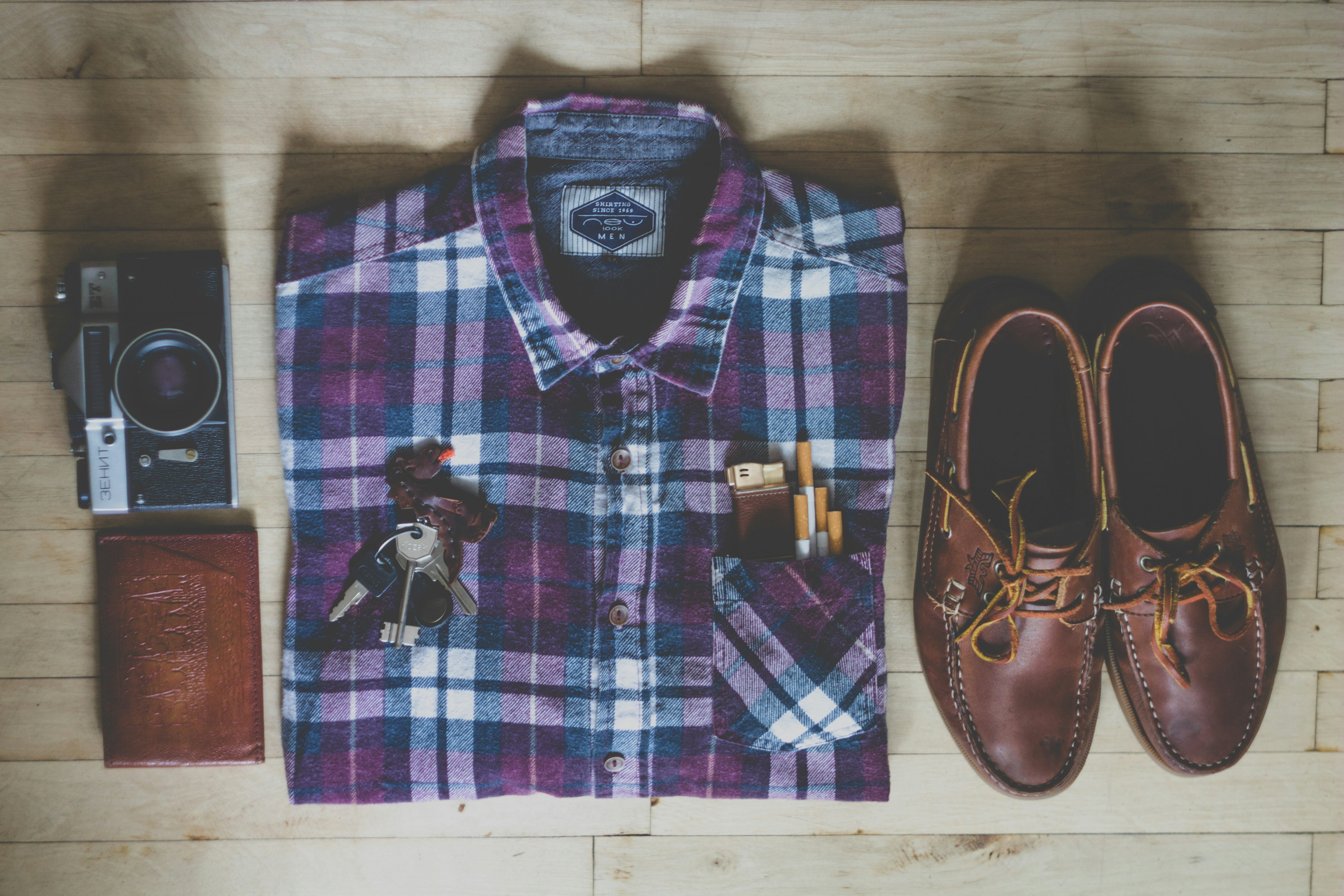 flat lay photography of button-up shirt, camera, keys, cigarettes, and pair of brown leather boat shoes